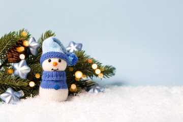 Knitted snowman near the branch of the Christmas tree with golden twinkling lights and decorations with blue stars on a blue background. Merry Christmas and New Year 2023 greeting card