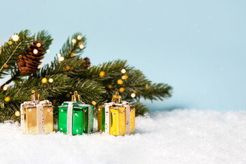 Three Christmas decorations in the form of gold and green gifts near the Christmas tree with twinkling golden lights on a blue background. Merry Christmas and New Year 2023 greeting card