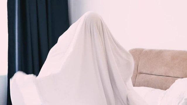A child in a sheet plays ghost in the bedroom on the bed. halloween. 