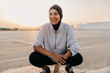 Happy Muslim sportswoman smiling while squatting outdoors