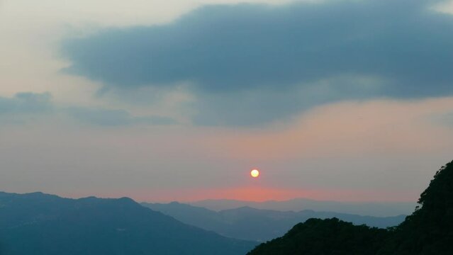 The sun sets behind the cascading hills. Tiny red sun. Orange-red sunset sky with fluffy clouds. Xindian District New Taipei City, Taiwan
