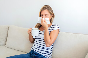 Young woman with cold blowing her runny nose. Unwell girl sneezing in a tissue in the living room. Sick young woman sneezing into a paper tissue while holding hot tea in domestic interior.