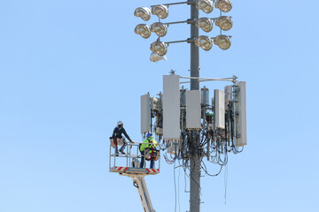 Workers using aerial lift maintain cell phone tower looming over high school football field