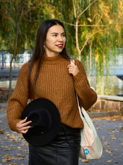 Attractive stylish woman holding hat and bag in hand, walking in park, dressed in warm suit. Pretty woman with brown hair smiles broadly teeth. Autumn fashion accessories in street fashion style