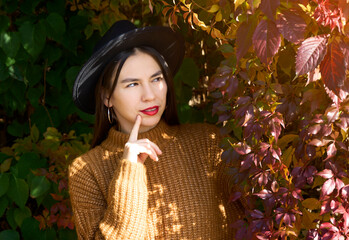 Selective focus of sweet dreamy young woman dressed brown outfit and hat. Finger on chin and thinking about autumn while standing in red and yellow leafs tree branches.