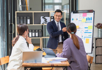 Team business three smiling and happy Asian men and women team using tablets and laptops brainstorm and plot statistics on a corporate chart board inside the company. Colleagues concept.