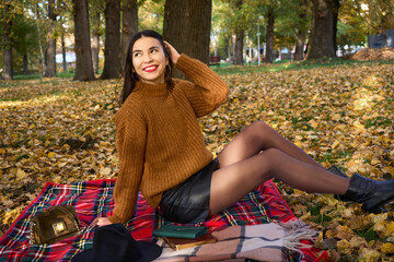 Fall picnic on october. Young woman with black hair and short leather skirt resting in nature on blanket and smiling. Autumn mood. People, lifestyle and relaxation.