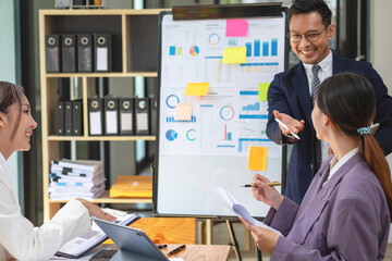 Team business three smiling and happy Asian men and women team using tablets and laptops brainstorm and plot statistics on a corporate chart board inside the company. Colleagues concept.