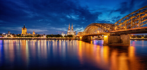 Panorama of illuminated bridge over Rhein river whith trains and tourists passing by and the Cologne Cathedral in the evening at the blue hour. Germany city life - obrazy, fototapety, plakaty