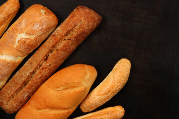 Bakery products on the background of a wooden dark table, top view. Baguettes, sesame buns and long loaf, fresh fragrant, crispy pastries