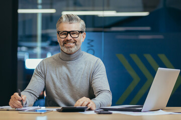 Portrait of successful senior businessman, gray haired man in glasses smiling and looking at camera, mature investor boss working inside modern office building using laptop at work.