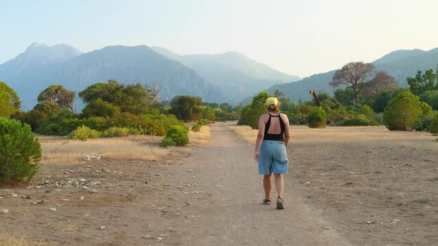 Girl in shorts, swimsuit and baseball cap walking the sandy path. Back view. Green pines and mountains in the background. Turkish savannah in the evening on the Lycian Trail