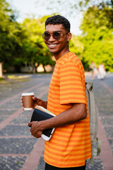 Fototapeta premium Young african man student holding coffee and books while standing outdoors