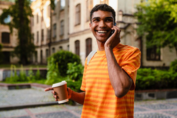 African man student talking on cellphone and drinking coffee while standing outdoors