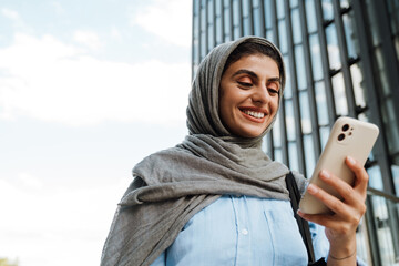 Muslim woman using smartphone while standing outdoors with glass building on background