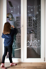 Little caucasian child girl decorating home window for Christmas with white pencil (liquid chalk). Festive home decor idea. Vertical shot.