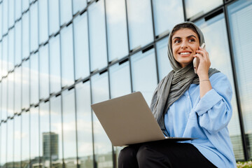 Cheerful muslim woman talking on cellphone and using laptop while sitting outdoors