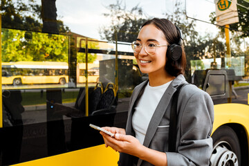 Young smiling asian woman holding smartphone and looking aside while standing near the bus outdoors