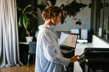 Businesswoman holding laptop and pressing button on panel of printer in office