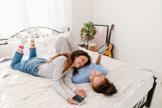 Two girls laughing and holding mobile phones while lying in bed together - Powered by Adobe