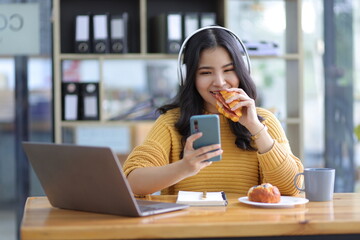 Attractive beautiful Asian woman casually working on smartphone in office and having snacks.