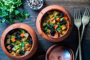 Stewed potato, carrot, onion, tomato and prunes in a clay pots with lid on wooden background, closeup