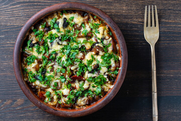 Stewed potato, carrot, onion, tomato, prunes and cheese in a clay bowl on wooden background, closeup, top view