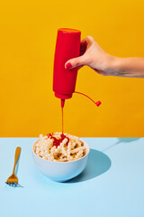Female hand pouring ketchup into bowl with necklaces symbolizing pasta, noodles on blue tablecloth over vivid yellow background. Food pop art photography.