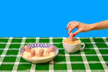 Plate with mashed potatoes and sausages on green tablecloth over blue background. Woman dipping sausage into coffee. Food pop art photography.