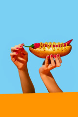 Female hands holding spicy chili hotdog with mustard over yellow tablecloth on blue background. Pepper instead of sausage. Food pop art photography.