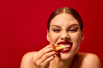 Emotive young woman eating fried potato, fries over vivid red background. Junk food lover. Delicious taste. Food pop art photography.
