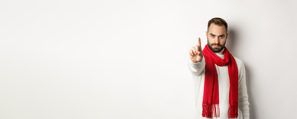 Serious man saying no, showing stop sign, shaking finger in decline, rejection sign, prohibit action, standing in winter sweater and red scarf against white background