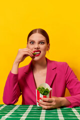 Emotional young woman in bright pink jacket eating green beans from fries packaging over yellow background. Healthy diet. Food pop art photography.