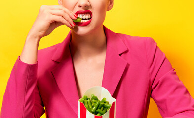 Cropped image of young woman in bright pink jacket eating green beans over yellow background. Dieting. Food pop art photography.