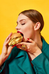 Young beautiful woman in green coat eating, biting cheeseburger with necklaces over yellow...