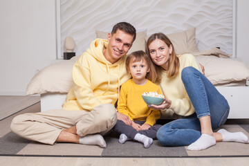 Friendly family, lovely parents in yellow clothes with child son sitting by the bed and eating popcorn, looking to the camera