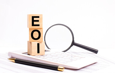 On a white calculator, next to a magnifying glass and a black pen, wooden cubes marked EOI...