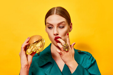 Young beautiful woman in green coat with red lipstick eating cheeseburger with necklaces, licking fingers over yellow background. Food pop art photography.
