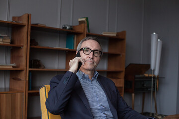 Portrait of elderly worried grey haired businessman in blue suit and eye glasses talking on mobile phone on grey studio background with bookshelves