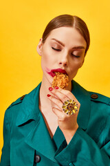 Young woman in green coat with red lipstick smudge, eating fried chicken, nuggets over yellow background. Food pop art photography.