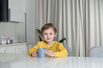 Happy cute child boy in yellow sweater sitting by the table and holding mug on white curtain background