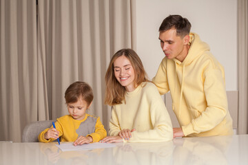 Fototapeta premium Adorable family, cute parents in yellow clothes with their child son sitting by the table and drawing and laughing