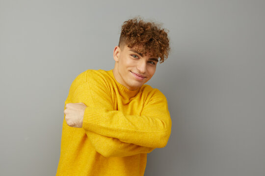 a nice young man stands on a gray background in a yellow sweater and hugging himself looks at the camera. Horizontal Studio Photography