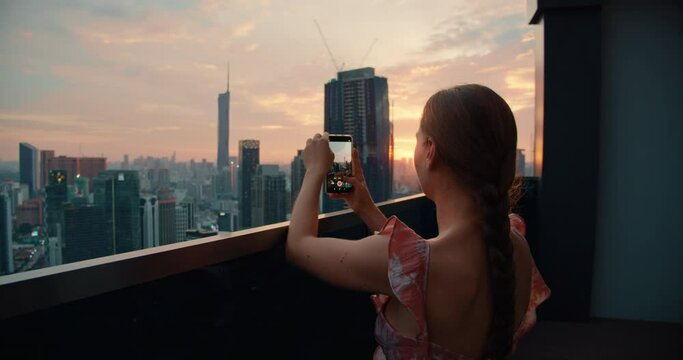 Woman on roof of skyscraper shoots a city landscape on a mobile phone during sunset. Rear view of tourist girl in twiligh on vacation. Kuala Lumpur capital of Malaysia.