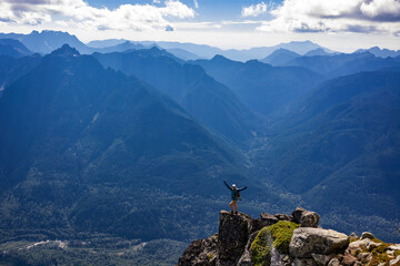 Fototapeta na wymiar Adventurous athletic female hiker standing on top of a cliff looking at the camera smiling with her arms held up in triumph with jagged mountains in the background in the Pacific Northwest.