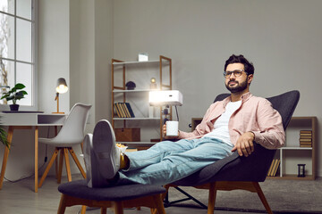 Happy relaxed young man sitting in a comfortable armchair at home, using a projector, watching a good movie, drinking coffee and enjoying quiet leisure time