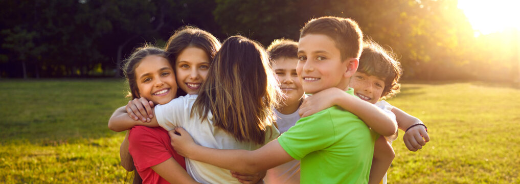Happy friends hugging in green summer park. Group of children having fun together. Bunch of cheerful little kids smiling and hugging each other tight. Outdoor shot with sun flare. Banner background