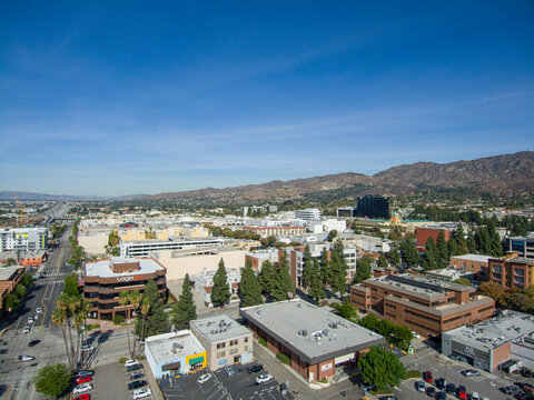 aerial shot of office buildings, apartments and shops surrounded by lush green trees, cars driving and parked on the street with majestic mountains and a clear blue sky in Burbank California USA