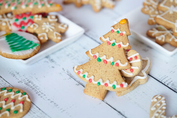 Galletas de gengibre de árbol navideño en mesa de madera blanca acompañada de más galletas con otros diseños en bandejas blancas.