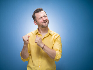 Amused handsome young man with closed eyes and clenched fists over blue background, dresses in yellow shirt. Yes concept. Amusing guy celebrates success